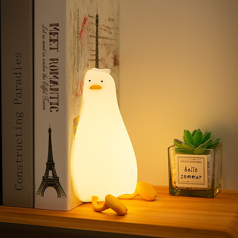 La veilleuse canard Ducky allumée, placée sur une étagère à côté d'un livre, créant une ambiance chaleureuse et accueillante.