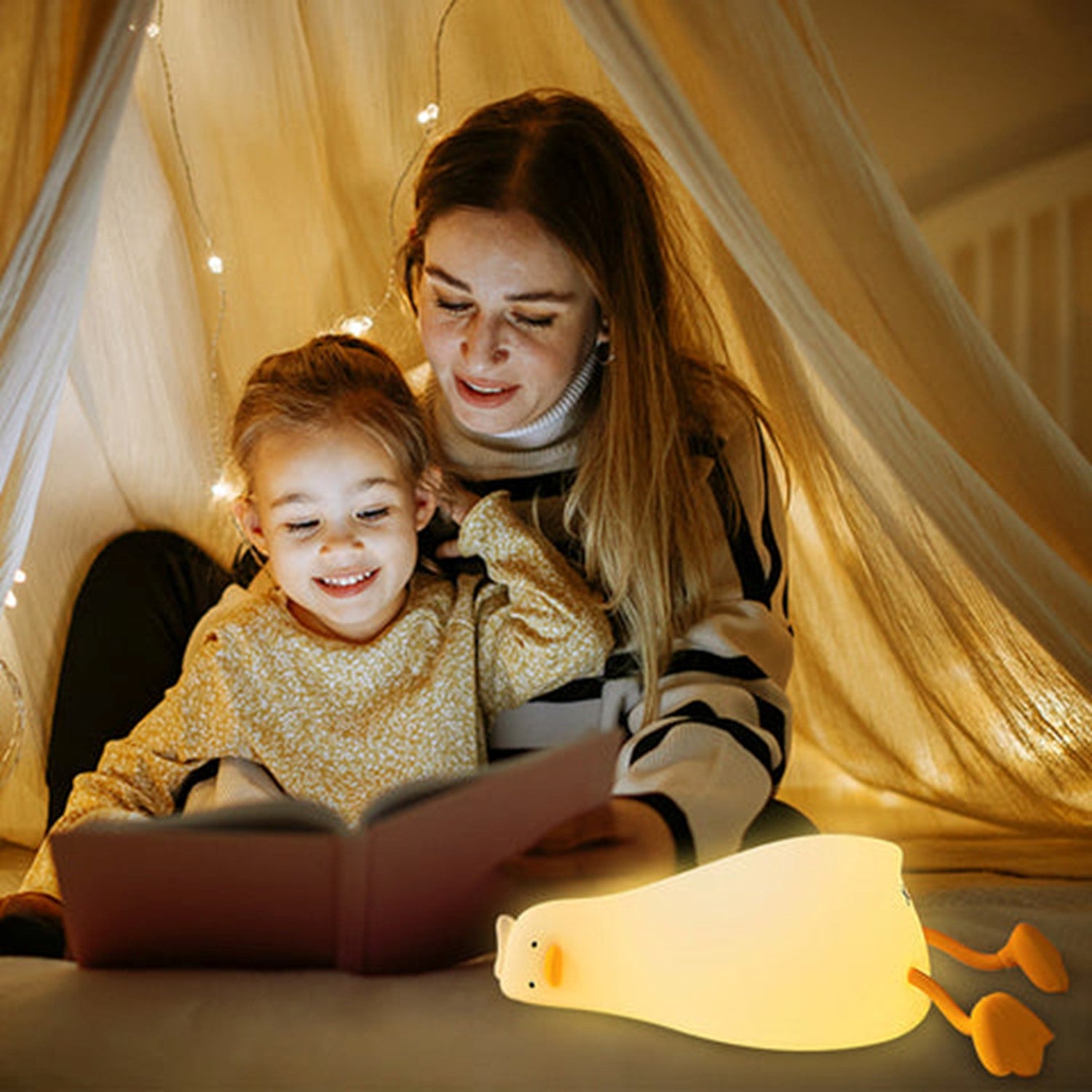 Veilleuse canard Ducky allumée illuminant une maman lisant un livre avec son enfant sous un ciel de lit, créant une ambiance magique. L'image dépeint la douceur et l'atmosphère enchantée que la veilleuse apporte lors des moments de lecture avant le coucher.