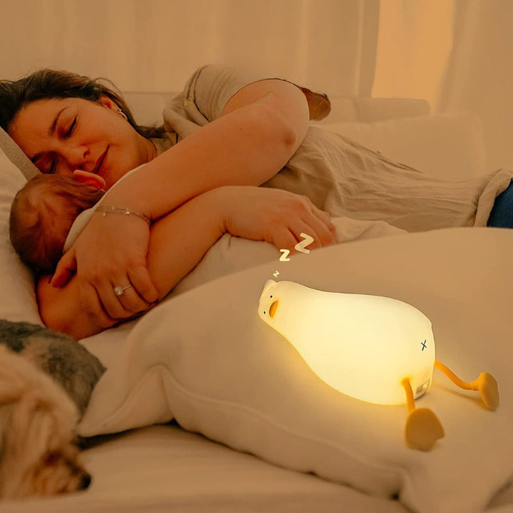 Veilleuse canard Ducky allumée placée sur un lit, à côté d'une maman tenant fermement son bébé dans ses bras, tous deux dormant paisiblement. L'image illustre le confort et la sécurité que la veilleuse apporte pendant le sommeil.